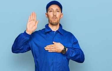 Kel, sakallı, inşaatçı üniforması giyen, elleri göğsünde ve avuç içi açık yemin eden, sadakat yemini eden bir adam. 