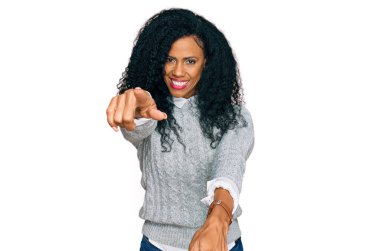 Orta yaşlı Afro-Amerikalı kadın günlük giysiler giyip seni ve kamerayı işaret ediyor, parmaklarıyla gülümsüyor ve neşeli. 