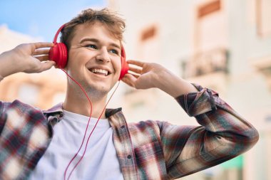 Genç beyaz adam şehirde kulaklık takarak mutlu bir şekilde gülümsüyor..