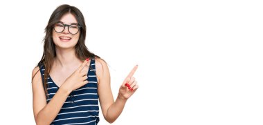 Gündelik kıyafetler giyen, gözlüklü, gülümseyen ve kameraya bakıp iki elini ve parmağını yan tarafa gösteren güzel beyaz bir kız.. 