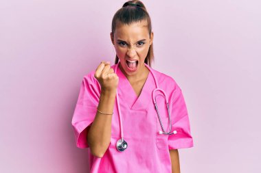 Genç İspanyol kadın doktor üniforması giyiyor ve steteskop kızgın ve kızgın yumruğunu kaldırıyor öfkeyle bağırırken sinirli ve öfkeli. öfke ve agresif konsept. 