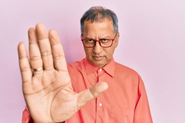 Orta yaşlı Hintli bir adam günlük kıyafetler ve gözlük takıyor. Avuç içiyle şarkı söylemeyi kesiyor. Yüzünde olumsuz ve ciddi bir jest olan uyarı ifadesi. 