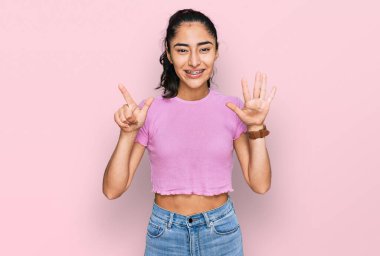 Diş teli takan, günlük kıyafetler giyen İspanyol genç kız yedi numaralı parmaklarıyla gülümseyerek kendinden emin ve mutlu bir şekilde onu işaret ediyor.. 