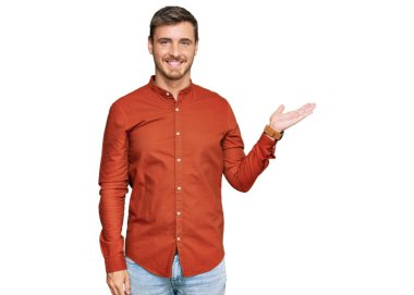 Yakışıklı, günlük kıyafetler giyen, neşeli bir şekilde gülümseyen, avuç içiyle kameraya bakan beyaz bir adam.. 