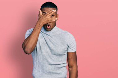 Gündelik tişört giyen genç siyah bir adam şok içinde yüzünü ve gözlerini kapayarak utangaç ifadesiyle parmaklarının arasından bakıyor.. 