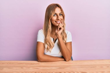 Güzel sarışın genç kadın, gündelik giysiler içinde masada oturmuş kameraya kendinden emin bir şekilde bakıyor kollarını kavuşturmuş ve çenesini kaldırmış bir şekilde gülümsüyor. Olumlu düşünmek. 