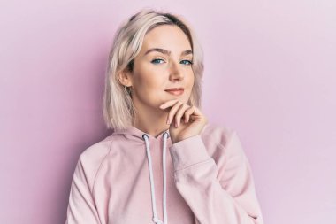 Gündelik kıyafetler giyen genç sarışın kız kameraya kollarını kavuşturup çenesine doğru bakıyor. Olumlu düşünmek. 