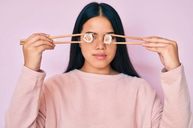 Genç, güzel Asyalı bir kız yemek çubuklarıyla gözlerine suşi sürüyor. Yüzünde ciddi bir ifade var. Basit ve doğal bir şekilde kameraya bakıyorsun.. 