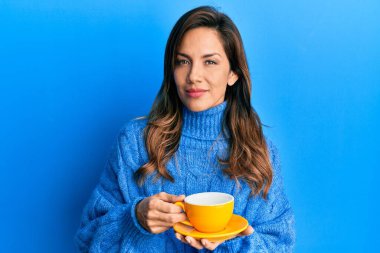 Bir fincan kahve içen genç Latin kadın yüzünde ciddi bir ifadeyle rahatlamış. Basit ve doğal bir şekilde kameraya bakıyorsun.. 