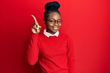 Genç Afro-Amerikalı kadın günlük kıyafetler ve gözlük takıyor. Bir numaralı parmağını gösterip işaret ederken kendinden emin ve mutlu gülümsüyor.. 