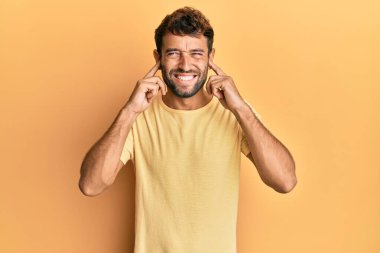 Sarı arka planda sarı tişört giyen sakallı yakışıklı adam yüksek sesli müzik sesi için kulaklarını parmaklarıyla kapatıyor. sağır kavramı. 