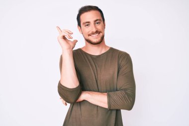 Yüzünde kocaman bir gülümseme olan sıradan kıyafetler giyen genç yakışıklı bir adam, kameraya el ve parmak ile bakıyor.. 