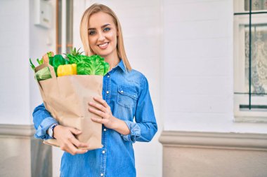 Genç sarışın kız gülümsüyor. Elinde alışveriş poşeti, şehirde duruyor..