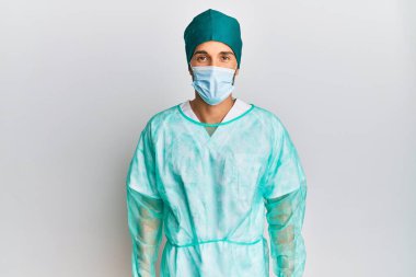 Young handsome man wearing surgeon uniform and medical mask sticking tongue out happy with funny expression. emotion concept. 