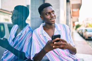 Afro-Amerikalı genç adam mutlu gülümsüyor. Şehrin caddesinde duvara yaslanmış akıllı telefon kullanıyor..