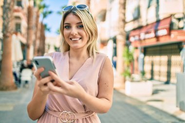 Genç sarışın kadın şehirde akıllı telefon kullanarak mutlu bir şekilde gülümsüyor..