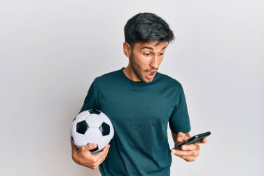 Young handsome man holding football ball looking at smartphone bet app celebrating crazy and amazed for success with open eyes screaming excited. 