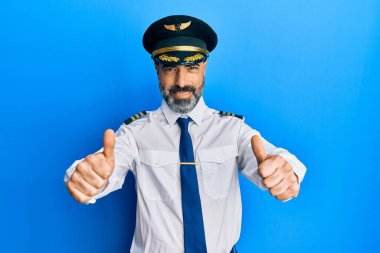 Orta yaşlı, sakallı, gri saçlı, uçak pilotu üniforması giyen, elle olumlu jest yapmayı onaylayan, gülümseyen ve başarı için mutlu bir adam. kazanan jest. 