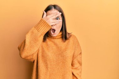 Young beautiful woman wearing turtleneck sweater peeking in shock covering face and eyes with hand, looking through fingers afraid 