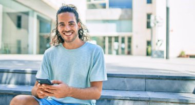 Genç İspanyol adam şehirde akıllı telefon kullanarak mutlu gülümsüyor..