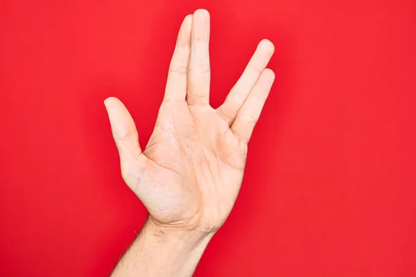 Hand of caucasian young man showing fingers over isolated red background greeting doing Vulcan salute, showing hand palm and fingers, freak culture