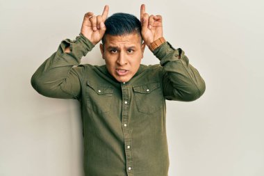 Gündelik kıyafetler giyen genç latin adam komik jestler yapıyor parmakları kafasının üstünde boğa boynuzu gibi 