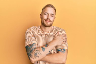 Kafkasyalı genç bir adam, günlük çizgili tişört giyiyor, kendine sarılıyor, mutlu ve pozitif, kendinden emin gülümsüyor. Sevgi ve özgüven. 