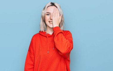 Genç sarışın kız gündelik kıyafetler giyiyor. Bir gözünü eliyle kapatıyor. Yüzünde kendine güvenen bir gülümseme ve sürpriz bir duygu var.. 