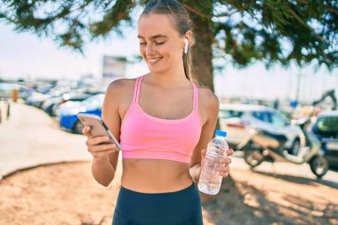 Genç sarışın bayan akıllı telefon kullanıyor ve şehirde bir şişe su içiyor..