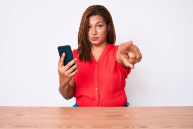 Orta yaşlı esmer kadın akıllı telefon kullanıyor masaya oturmuş kamerayı işaret ediyor ve sen kendine güvenen ciddi bir hareket yapıyorsun. 