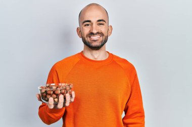 Elinde çiğ fındık tutan genç kel adam pozitif ve mutlu görünüyor. Durup gülümsüyor ve kendine güvenen bir gülümsemeyle dişlerini gösteriyor. 