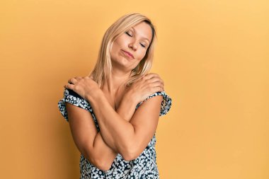 Orta yaşlı beyaz kadın günlük kıyafetler giyip kendine sarılıyor mutlu ve pozitif, kendinden emin gülümsüyor. Sevgi ve özgüven. 