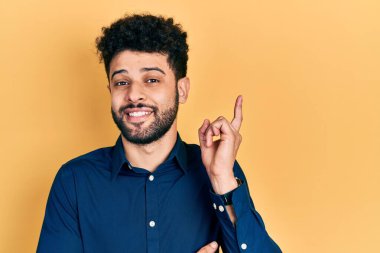 Sakallı, rahat gömlekli, yüzünde kocaman bir gülümseme olan genç bir Arap adam, kameraya el ve parmak ile bakıyor.. 