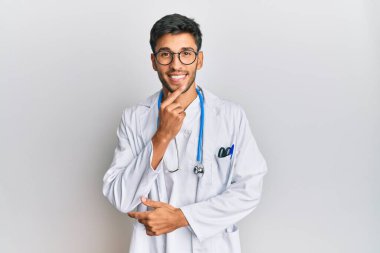 Young handsome man wearing doctor uniform and stethoscope looking confident at the camera with smile with crossed arms and hand raised on chin. thinking positive. 