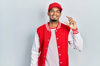 Beyzbol forması giyen, gülümseyen, kendine güvenen, el hareketi yapan, parmaklarıyla işaret yapan ve kameraya bakan Afrikalı genç bir Amerikalı. ölçüm kavramı. 