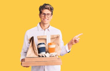 İş kıyafetleri içinde genç yakışıklı bir adam elinde teslimat yemeği tutarken mutlu bir şekilde elini ve parmağını kenarda gezdirirken. 