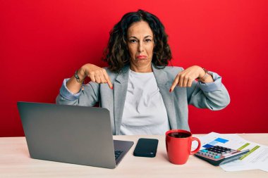 Ofiste çalışan orta yaşlı güzel bir kadın bir fincan kahve içiyor üzgün ve üzgün görünüyor, parmaklarıyla yönü gösteriyor, mutsuz ve depresif.. 