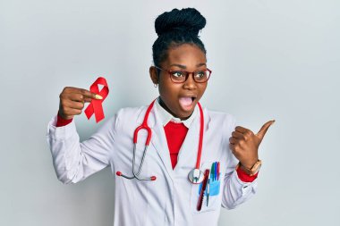 Genç Afro-Amerikan kadın doktor üniforması giyiyor. Elinde destek kırmızı kurdelesi var. Başparmağıyla yan tarafı işaret ediyor. 
