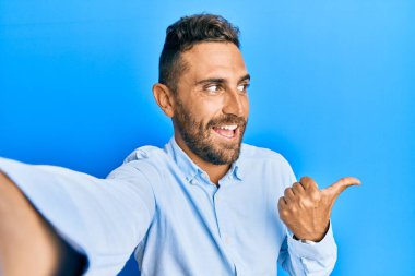 Sakallı yakışıklı bir adam akıllı telefonuyla fotoğraf çekiyor. Parmağını kaldırıyor ve ağzı açık gülümsüyor. 