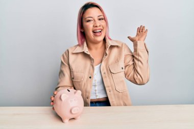 Pembe saçlı İspanyol kadın domuz kumbarasıyla başarıyı kutluyor. Mutlu bir gülümseme ve kaldırılmış bir el ile kazanan yüz ifadesi. 