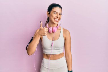 Genç İspanyol kadın spor kıyafetleri giyiyor ve kulaklık takıyor. Elleriyle mutlu bir jest yapıyor. Kameraya bakıp başarıyı gösteren ifade. 