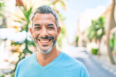 Orta yaşlı, kır saçlı, gülümseyen bir adam şehrin caddesinde mutlu bir şekilde yürüyor..