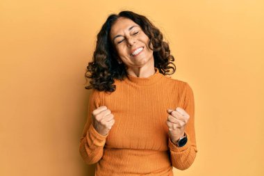 Orta yaşlı İspanyol kadın gündelik giysiler giyiyor başarı için heyecanlı kolları havada ve gözleri kapalı zaferi kutlarken gülümsüyor. Kazanan konsept. 