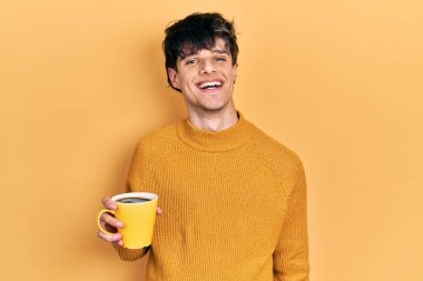 Yakışıklı hippi genç adam kahve içiyor pozitif ve mutlu görünüyor ve gülümsüyor kendine güvenen bir gülümsemeyle dişlerini gösteriyor. 