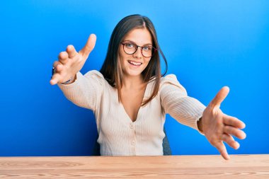 Gündelik giysiler ve gözlük takmış genç beyaz bir kadın kucaklanmak için kollarını açmış kameraya bakıyor. Mutluluğu kucaklayan neşeli bir ifade.. 
