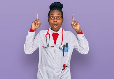 Genç Afro-Amerikan kadın doktor üniforması ve steteskop giyiyor. Şaşırmış ve şaşırmış bir şekilde yukarı bakıyor ve parmaklarıyla işaret ediyor ve kollarını kaldırıyor.. 
