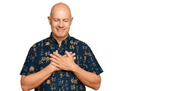 Orta yaşlı kel bir adam, sıradan kıyafetler giyiyor, elleri göğsünde, gözleri kapalı gülümsüyor ve yüzünde minnet ifadesi var. sağlık kavramı. 