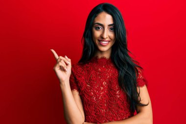Güzel İspanyol kadın, kırmızı arka planda zarif kıyafetler giyiyor. Gülümsüyor, mutlu bir şekilde elini ve parmağını yana doğru gösteriyor. 