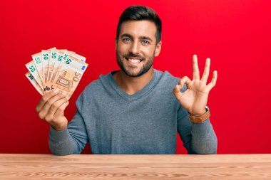 50 Euro 'luk banknotlar tutan yakışıklı İspanyol adam parmaklarıyla imza atıyor, gülümseyerek mükemmel bir sembol oluşturuyor. 