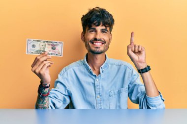 50 dolarlık banknotu elinde tutan genç İspanyol bir adam masaya oturmuş gülümsüyor bir fikir veya soru işareti ile mutlu bir yüzle, bir numara 